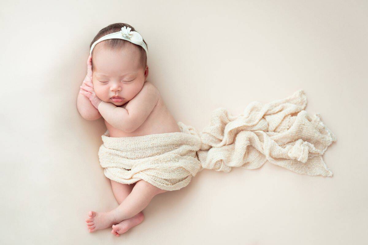 Newborn baby girl wrapped in a cream blanket with a delicate white flower headband — perfect for a post on elegant girl names.