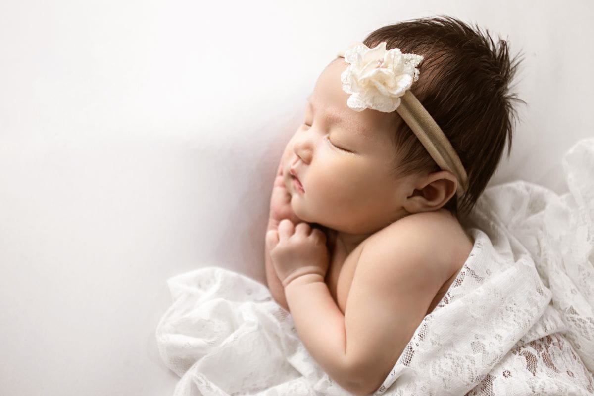 Newborn baby girl sleeping peacefully on a white blanket, wearing a lace headband with a cream flower and wrapped in delicate lace fabric.