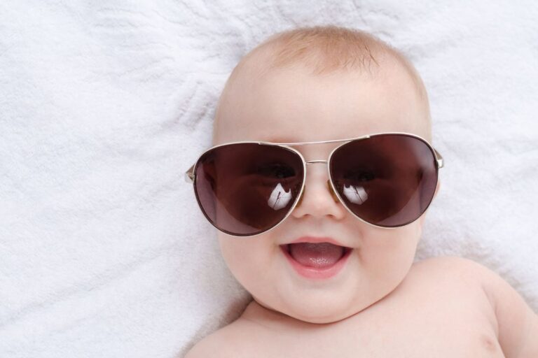 A smiling baby lying on a soft white towel wearing oversized aviator sunglasses, giving off a cool and confident vibe – perfect for a post about edgy boy names.